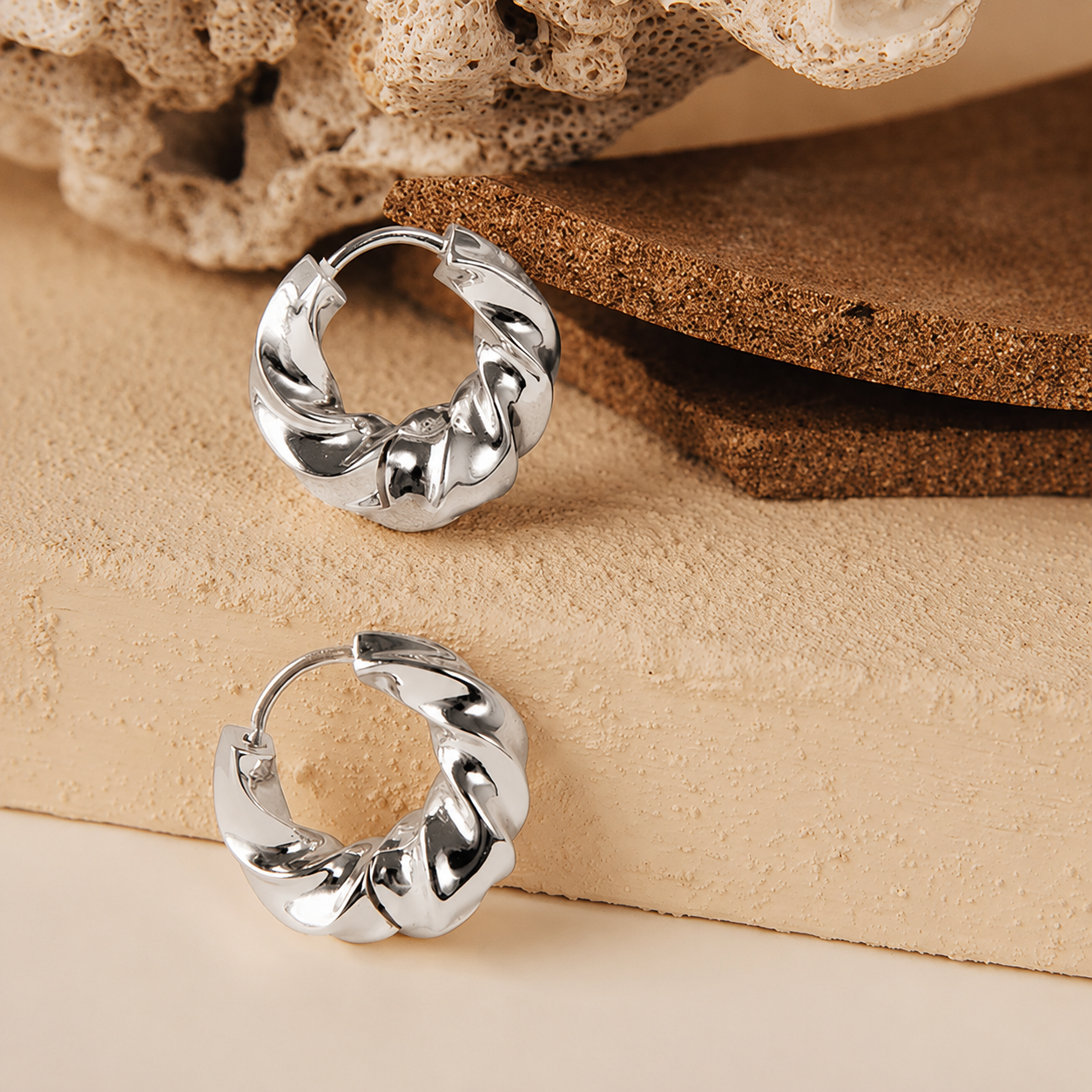 A pair of polished silver hoop earrings with a twisted, textured design, displayed on a beige textured surface with a cork and bone prop.